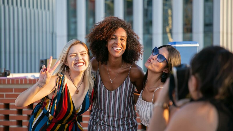 Unrecognizable female photographer taking picture of cheerful young multiracial female friends smiling and showing V sign during party on modern building rooftop